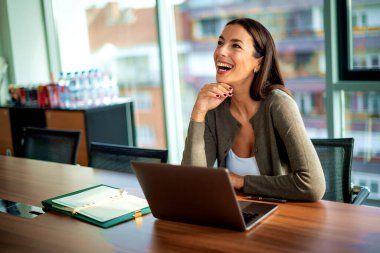 İş için bilgisayarını kullanan gülümseyen bir iş kadını. Çekici profesyonel kadın iş kıyafeti giyiyor ve modern bir ofiste oturuyor.. 