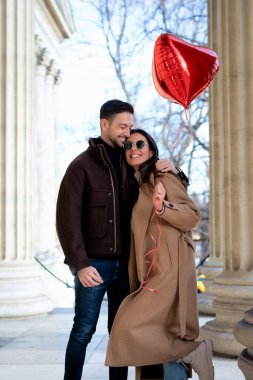 Şehirde yürüyüp eğlenen mutlu bir çift. Kafkasyalı kadın kalp şeklinde balon tutuyor ve sevgililer gününü kutluyor.. 