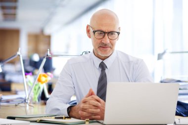 Ofisindeki masasında oturan orta yaşlı bir işadamı laptopuyla çalışıyor. Tişört ve kravat takan kendine güvenen profesyonel bir adam ve defterine özenle bakıyor..  