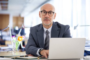 Ofisindeki masasında oturan orta yaşlı bir işadamı laptopuyla çalışıyor. Kendine güvenen, takım elbiseli, kravatlı ve düşünceli bir adam..  