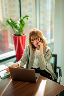 İş için bilgisayarını kullanan gülümseyen bir iş kadını. Çekici profesyonel kadın iş kıyafeti giyiyor ve modern bir ofiste oturuyor.. 