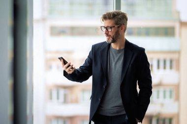 Modern bir ofiste duran ve akıllı telefonunu kullanan bir iş adamı. İş kıyafeti giyen kendine güvenen bir adam..