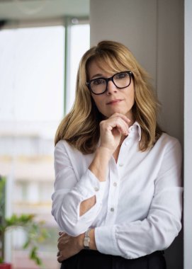 Cam duvarda duran sarı saçlı kadın. Beyaz gömlek giyen ve okuma gözlüğü takan orta yaşlı bir kadın..