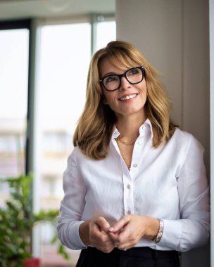 Cam duvarda duran sarı saçlı kadın. Beyaz gömlek giyen ve okuma gözlüğü takan orta yaşlı bir kadın..