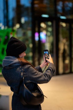 Sokakta dikilip akıllı telefonunu kullanan çekici bir kadın. Bir sonbahar gününde şapka ve eşarp takan mutlu bir kadın. Arka plan görüntüsü.