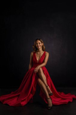 Kırmızı elbise giyen çekici orta yaşlı kadın. Sarışın kadın kameraya bakıyor ve izole edilmiş karanlık arka planda gülümsüyor.. 