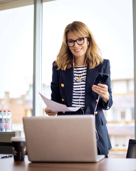 Деловая женщина средних лет, стоящая за своим столом с наушниками и сотовым телефоном. Уверенная женщина в пиджаке и очках для чтения. 