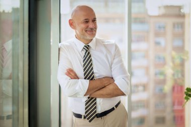 Modern bir ofiste kolları katlanmış, pencerenin kenarında duran orta yaşlı bir adam. Beyaz iş adamı gömlek ve kravat giyiyor.. 