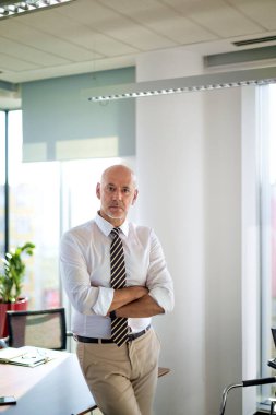 Modern bir ofiste ofis masasında yönetici bir işadamı duruyor. Gömlek ve kravat takıp kameraya bakan orta yaşlı profesyonel bir adam..