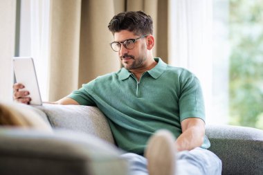 Orta yaşlı bir adam evdeki kanepede dinleniyor ve touhcpad 'ini kullanıyor. Avrupalı erkek günlük kıyafetler giyiyor ve okuma gözlüğü takıyor. 