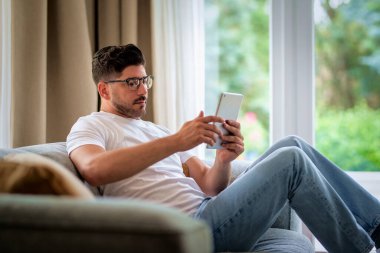 Orta yaşlı bir adam evdeki kanepede dinleniyor ve touhcpad 'ini kullanıyor. Avrupalı erkek günlük kıyafetler giyiyor ve okuma gözlüğü takıyor. 