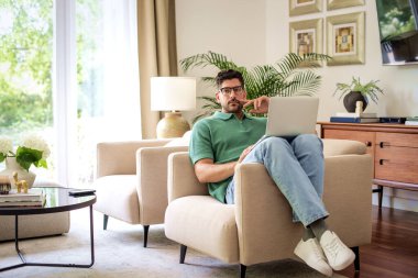 Orta yaşlı bir adam evinde koltuğunda dizüstü bilgisayarını kullanıyor. Avrupalı erkek, evden çalışıyor. Merkez ofis. Tam boy çekim. 