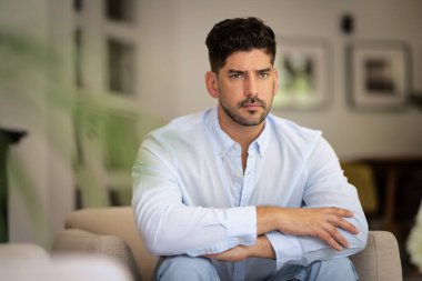 Sakallı, orta yaşlı bir adam evinde koltuğunda oturuyor ve düşünceli görünüyor. Gündelik kıyafetler giyen kendinden emin bir erkek.. 