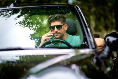 Güneş gözlüğü takan, üstü açık bir arabada oturan ve yolda araba süren yakışıklı bir adam. Akıllı telefonunu kullanan orta yaşlı bir erkek.. 