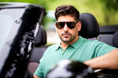 Güneş gözlüğü takan, üstü açık bir arabada oturan ve yolda araba süren yakışıklı bir adam.. 