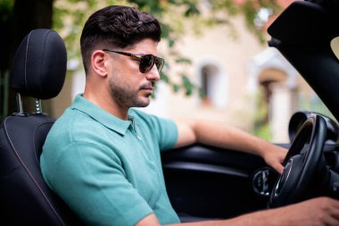 Güneş gözlüğü takan, üstü açık bir arabada oturan ve yolda araba süren yakışıklı bir adam.. 