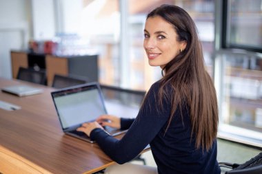 Ofis masasında dizüstü bilgisayarının arkasında oturmuş gülümseyen bir iş kadını. Çekici profesyonel kadın iş kıyafeti giyiyor ve kameraya bakıyor.. 