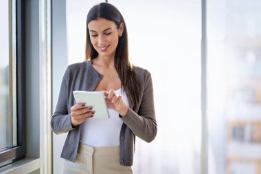 İş kadını ofiste dijital tabletini kullanıyor. Kendine güveni olan, sıradan kıyafetler giyen ve neşeli gülümseyen profesyonel bir kadın..