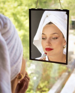 Güzel genç bir kadın kırmızı ruj sürerken pencerede dikilip aynaya bakıyor..