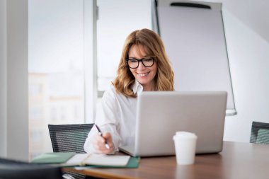 Kendine güvenen olgun bir iş kadını modern bir ofiste masa başında oturuyor ve bilgisayarını kullanıyor. Profesyonel bir kadın konferans görüşmesi yapıyor.. 