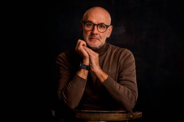 Karanlık bir arkaplanda dalgın dalgın oturan son sınıf öğrencisi. Okuma gözlüğü ve süveter giyen kendinden emin bir erkek. İzole edilmiş arka plan. Boşluğu kopyala. 