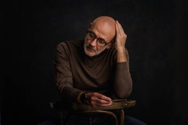 Karanlık bir arkaplanda dalgın dalgın oturan son sınıf öğrencisi. Okuma gözlüğü ve süveter giyen kendinden emin bir erkek. İzole edilmiş arka plan. Boşluğu kopyala. 