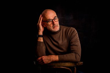 Karanlık bir arkaplanda dalgın dalgın oturan son sınıf öğrencisi. Okuma gözlüğü ve süveter giyen kendinden emin bir erkek. İzole edilmiş arka plan. Boşluğu kopyala. 