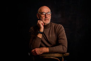 Karanlık bir arkaplanda dalgın dalgın oturan son sınıf öğrencisi. Okuma gözlüğü ve süveter giyen kendinden emin bir erkek. İzole edilmiş arka plan. Boşluğu kopyala. 