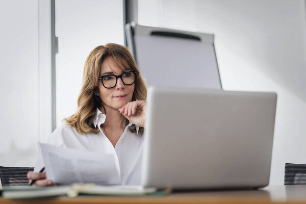 Уверенная зрелая деловая женщина, сидящая за столом в современном офисе и пользующаяся своим ноутбуком. Профессиональная женщина проводит конференцию. 