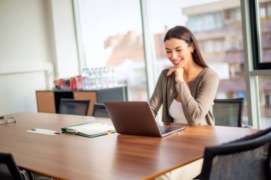 Ofis masasında dizüstü bilgisayarının arkasında oturmuş gülümseyen bir iş kadını. İş kıyafeti giyen çekici profesyonel kadın.. 