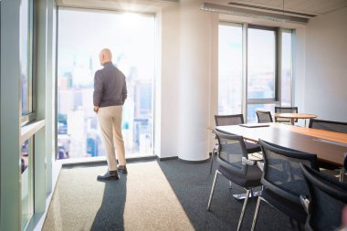 Ofise arkası dönük duran ve pencereden şehre bakan bir adam. Tam yol ileri. İş kıyafeti giyen profesyonel bir erkek.. 