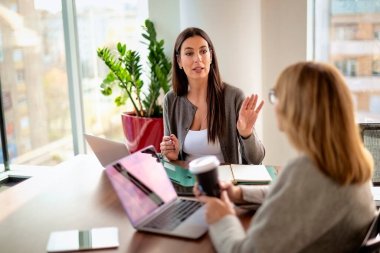Modern bir ofiste masa başında oturan ve dizüstü bilgisayarlarla çalışan iki iş kadını. Takım çalışması. Bir grup iş adamı toplantı sırasında tartışıyor.