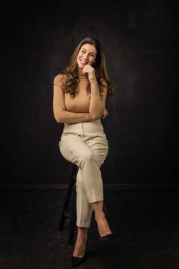 Esmer, neşeli, gülümseyen ve ıssız, karanlık arka planda oturan bir kadın. Balıkçı yaka kazak ve pantolon giyen çekici bir kadın. Tam yol ileri. Uzayı kopyala Elini yüzüne koy..