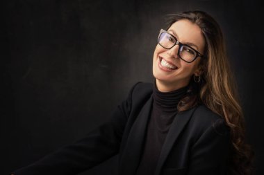 Esmer, neşeli, gülümseyen ve izole edilmiş karanlık arka planda oturan bir kadın. Balıkçı yaka kazak ve gözlük giyen çekici bir kadın.. 