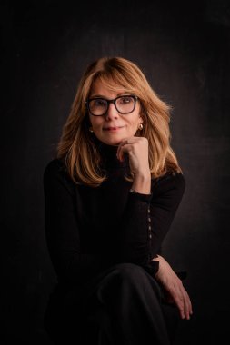 Sarışın, gülümseyen ve izole edilmiş karanlık arka planda oturan bir kadın. Balıkçı yaka kazak ve gözlük giyen çekici bir kadın. Uzayı kopyala Eller çenede.. 