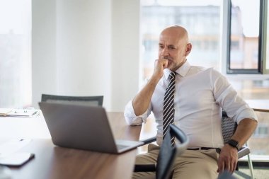 Ofis masasında dizüstü bilgisayarı olan beyaz bir adam oturuyor ve bir defter kullanıyor. Profesyonel işadamı düşünceli görünüyor ve web konferansı düzenliyor..