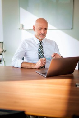 Ofis masasında dizüstü bilgisayarı olan beyaz bir adam oturuyor ve bir defter kullanıyor. Profesyonel işadamı düşünceli görünüyor ve web konferansı düzenliyor..