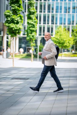 Gülümseyen orta yaşlı bir adam şehir sokaklarında yürüyor. Kendine güvenen iş adamı dizüstü bilgisayar tutuyor ve iş kıyafeti giyiyor.. 