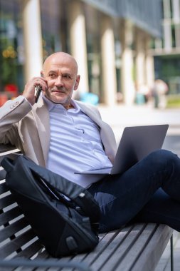 Şehirdeki bir bankta oturan yakışıklı iş adamı cep telefonu ve dizüstü bilgisayar kullanıyor. Profesyonel erkek iş kıyafeti giyiyor ve çalışıyor.. 