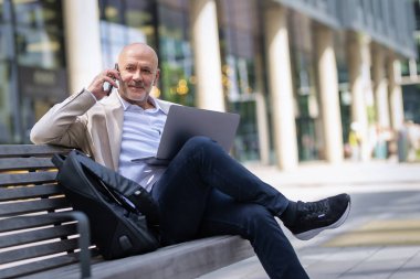 Şehirdeki bir bankta oturan yakışıklı iş adamı cep telefonu ve dizüstü bilgisayar kullanıyor. Profesyonel erkek iş kıyafeti giyiyor ve çalışıyor.. 