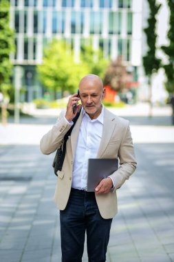 Yakışıklı bir adam dizüstü bilgisayar tutuyor ve şehirde finans sektöründe yürüyor. Orta yaşlı işadamı iş kıyafeti giyiyor ve telefon görüşmesi yapıyor.. 