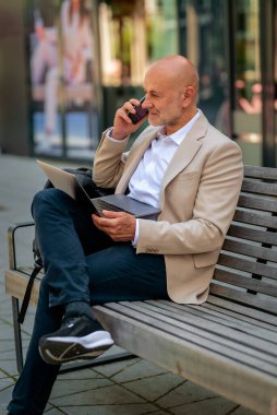 Şehirdeki bir bankta oturan yakışıklı iş adamı cep telefonu ve dizüstü bilgisayar kullanıyor. Profesyonel erkek iş kıyafeti giyiyor ve çalışıyor. Tam boy çekim. 