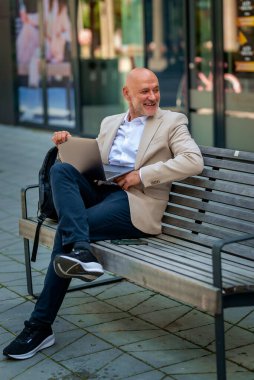 Şehirde bir bankta oturan yakışıklı bir iş adamı ve dizüstü bilgisayar kullanıyor. Profesyonel erkek iş kıyafeti giyiyor ve çalışıyor. Tam boy çekim.