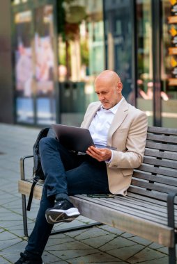 Şehirde bir bankta oturan yakışıklı bir iş adamı ve dizüstü bilgisayar kullanıyor. Profesyonel erkek iş kıyafeti giyiyor ve çalışıyor. Tam boy çekim.