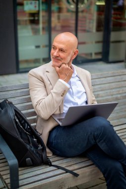 Şehirde bir bankta oturan yakışıklı bir iş adamı ve dizüstü bilgisayar kullanıyor. Profesyonel erkek iş kıyafeti giyiyor ve çalışıyor..