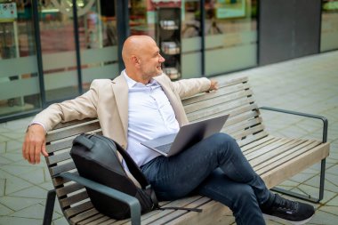 Şehirde bir bankta oturan yakışıklı bir iş adamı ve dizüstü bilgisayar kullanıyor. Profesyonel erkek iş kıyafeti giyiyor ve çalışıyor..