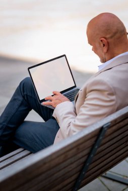 Şehirdeki bir bankta oturan ve dizüstü bilgisayar kullanan bir iş adamının yakın çekimi. Profesyonel erkek iş kıyafeti giyiyor ve çalışıyor. Boş ekran. 