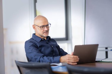 Orta yaşlı profesyonel bir adam modern bir ofiste masa başında oturmuş web konferansı yapıyor. Kendine güvenen işadamı iş kıyafeti giyiyor ve iş için laptop kullanıyor.. 