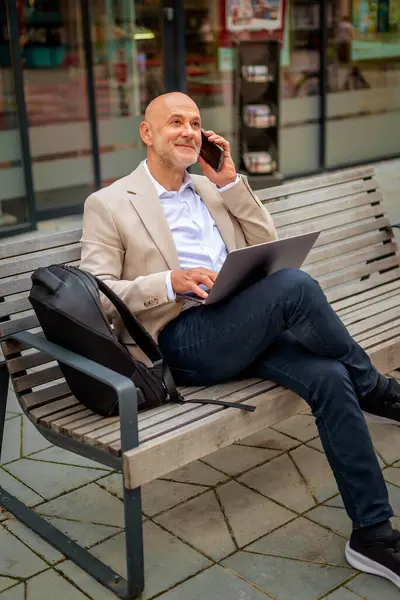 Şehirdeki bir bankta oturan yakışıklı iş adamı cep telefonu ve dizüstü bilgisayar kullanıyor. Profesyonel erkek iş kıyafeti giyiyor ve çalışıyor. Tam boy çekim. 