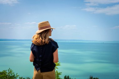 Denize bakan bir kadının dikiz görüntüsü. İlham ve farkındalık kavramı. Hasır şapka takan ve sıradan kıyafetler giyen bir kadın..